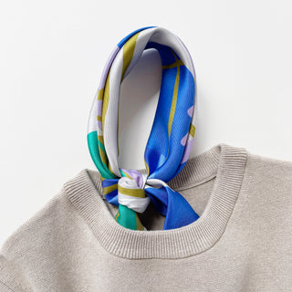 Abstract lotus silk scarf in vibrant blue, green, lilac, and yellow patterns tied and tucked in a beige sweater
