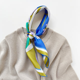 Abstract lotus silk scarf in vibrant blue, green, lilac, and yellow patterns tied as a neckerchief on a beige sweater