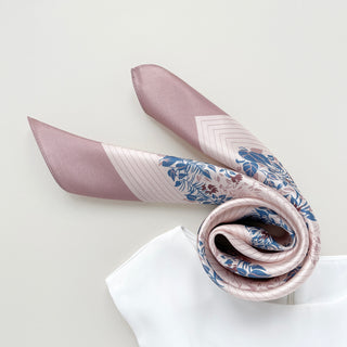 Blush pink silk scarf with blue and burgundy leaf pattern elegantly rolled, displaying its shimmery texture and decorative border design on white background.