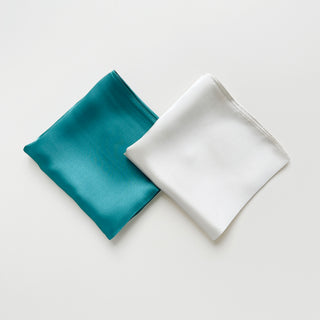 Two folded square silk scarves, one teal and one white, on a light grey background