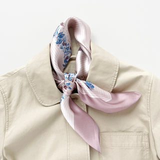 Blush pink silk bandana scarf with blue and burgundy leaf print, elegantly tied around collar of beige jacket, showing its luxurious sheen.