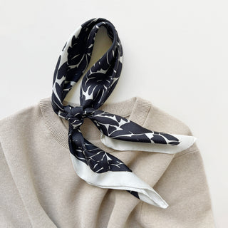 Luxury silk scarf in black floral print with ice white border draped over a beige sweater on a light grey background