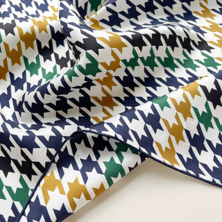 Close-up of a luxury silk scarf featuring a houndstooth pattern in navy, green, and gold on white background, with elegant hand-rolled edges and smooth fabric texture.