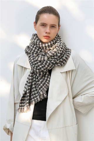 Woman wearing a taupe, black and cream white cashmere scarf with houndstooth check pattern and white coat against a light background