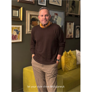 Brown and orange English plaid cashmere silk scarf styled with brown sweater and patterned pants, displayed in cozy home setting with yellow sofa.