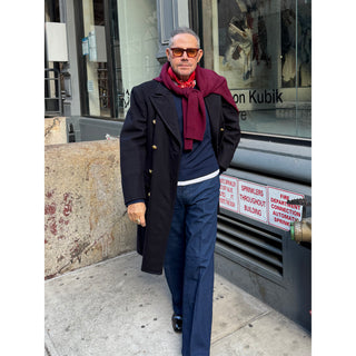 Man wearing a red paisley silk scarf by LuluLane in a dark coat and sunglasses walking on a sidewalk with a store window in the background.