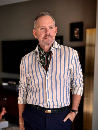 Men’s silk and cotton neck scarf in rich green with stylised bird print, styled with a white and blue striped shirt, navy trousers.