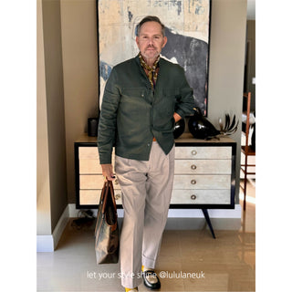 Men’s silk and cotton neck scarf in olive with stylised bird print, styled with a dark green jacket, pleated trousers, and a leather tote.