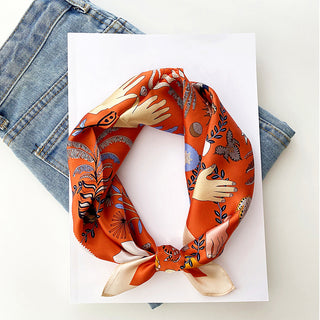 Orange silk bandana scarf with mystical tarot designs featuring hands, plants and symbols, displayed on white paper against denim