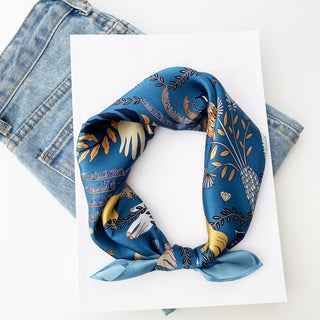 Dusty blue silk bandana scarf with mystical tarot-inspired design featuring gold leaves, moons, and hands on white paper against denim