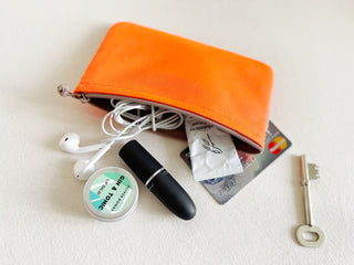 Bright orange recycled zipper wallet with various everyday items displayed: earbuds, lip balm, credit card, and key on neutral background.