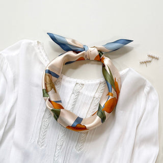 Blue Orange Iris Melody silk bandana scarf tied around white blouse collar, featuring floral and butterfly print in blue, orange, and cream tones