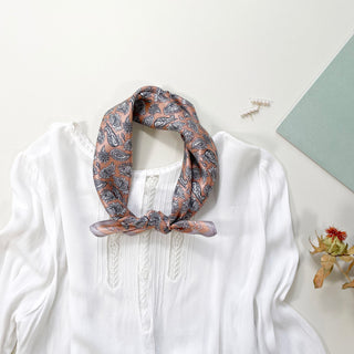 Pink silk bandana with navy paisley pattern tied as a neckerchief on white blouse, styled with pearl hairpin and minimalist decor.
