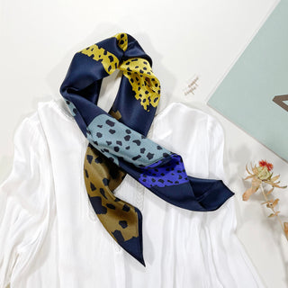 Square silk scarf with navy blue border and leopard pattern in yellow, blue, and bronze tones, draped on a white blouse with dried flower accent.