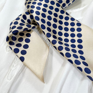 Silk bandana scarf with navy blue polka dots on light beige background, displayed on white fabric showing its glossy texture and elegant pattern.