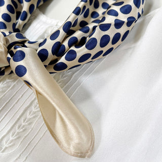 Silk bandana scarf with navy blue polka dots on light beige background, showing glossy texture and corner detail on white fabric