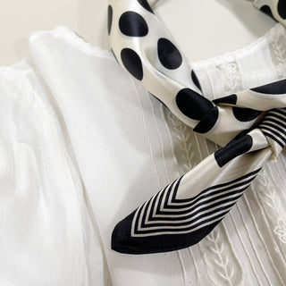 Luxurious cream silk bandana scarf featuring bold black polka dots and geometric striped border pattern, draped elegantly over white fabric.