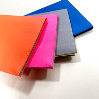 Four minimalist recycled passport holders in vibrant colors - orange, pink, gray, and blue - stacked with neat piping detail, made from RPET material