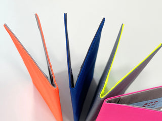 Four minimalist passport holders made from recycled RPET in vibrant colors: orange, blue, gray with neon yellow trim, and pink, arranged in a fan display.