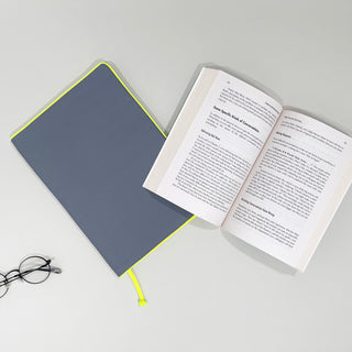 Eco-friendly notebook cover in navy blue with neon yellow trim, shown next to an open book and reading glasses on white background.