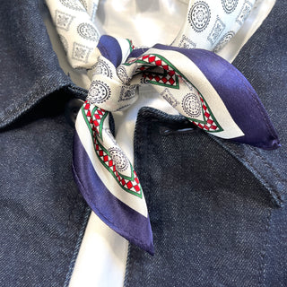 Men's silk neckerchief with cream and purple colors, featuring decorative medal patterns, red and green geometric borders, elegantly tied over a dark denim jacket.