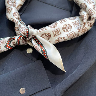 Cream silk neckerchief with brown medal pattern and decorative red-green border, elegantly knotted against navy shirt, showcasing its luxurious sheen