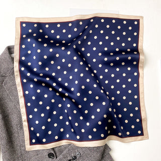 Navy blue silk square scarf with white polka dots pattern and cream-colored border, displayed on a textured fabric surface.