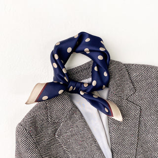 Navy blue silk scarf with cream polka dots tied as a neckerchief on a patterned blazer, showcasing the scarf's lustrous finish and elegant style