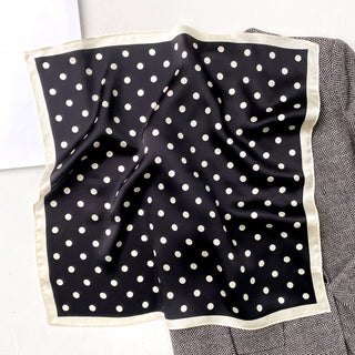 Black silk bandana scarf with white polka dots and cream border, displayed on patterned surface showing luxurious fabric texture