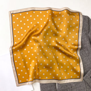 Mustard yellow silk bandana scarf with white polka dots and cream border, displayed on geometric patterned fabric showing its luxurious texture