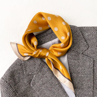 Mustard yellow silk bandana scarf with white polka dots tied elegantly around collar, contrasting against geometric patterned garment
