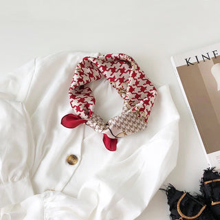 Red and white houndstooth silk bandana scarf elegantly tied around white blouse collar, displayed with fashion magazine and accessory
