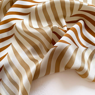 Luxurious 100% silk scarf with elegant golden caramel stripes on cream background, showing soft folds and lustrous sheen texture