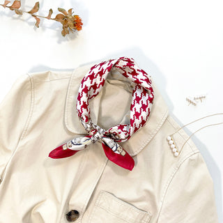 Red and white houndstooth silk bandana scarf tied elegantly around collar of beige jacket, with decorative pearl pin and dried flower accent