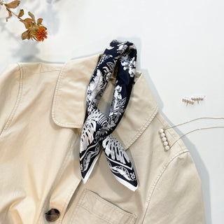Black silk bandana with white fern and monstera leaf pattern draped on beige jacket, styled with pearl accessories against soft background