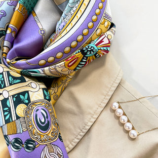 Luxurious silk scarf in yellow and lilac with vintage decorative pattern, draped elegantly over beige fabric alongside pearl necklace