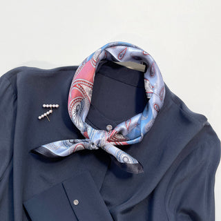 Salmon pink and light blue paisley silk bandana tied around collar of navy shirt with pearl brooch, showing elegant black-bordered scarf pattern