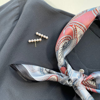 Salmon pink and light blue paisley silk scarf styled as a neckerchief on dark gray blazer with pearl brooch, showing intricate pattern and black border