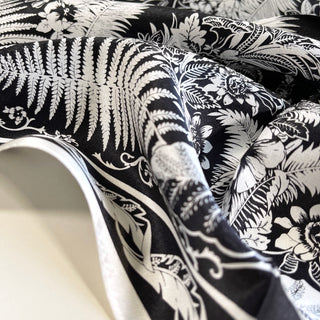 Black silk scarf with elegant white botanical pattern featuring detailed ferns, flowers, and monstera leaves in a luxurious rippled fabric texture