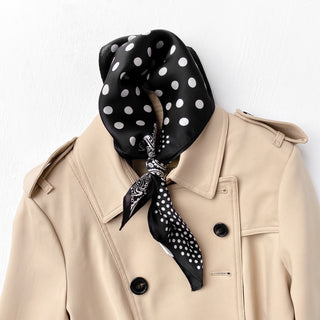 Black silk bandana with white polka dots and paisley pattern, elegantly tied around the collar of a beige trench coat