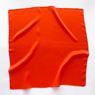 Square orange silk scarf with smooth, lustrous finish displayed flat with gentle folds showing the fabric's softness and light-reflecting quality