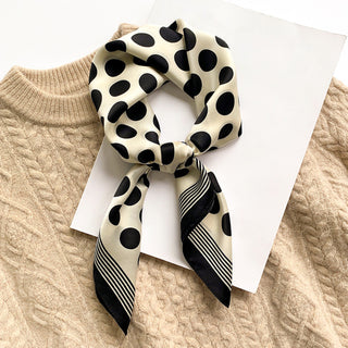 Silk bandana scarf with cream background and black polka dots, featuring striped border, styled in knot over beige cable knit sweater
