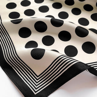 Luxurious silk scarf with black polka dots on cream background and elegant black striped border, showcasing silky fabric texture and classic pattern