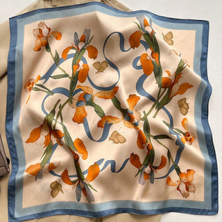 Luxurious silk bandana scarf with cream background featuring orange iris flowers, delicate butterflies, and flowing blue ribbon pattern, bordered in soft blue.