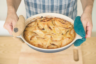 Hands holding a baked apple dessert using contrasting linen oven mitts - tan with embroidered detail on left, teal blue on right.