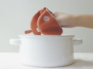 Terracotta orange linen oven mitt with white piping and embroidered detail, held over a white cooking pot, showcasing its compact size and stylish design