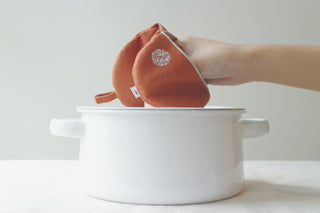Compact terracotta-colored linen oven mitt with white piping and embroidered circle detail being used to lift a white pot lid