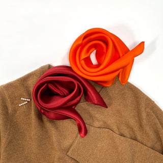 Two classic silk scarves in burgundy and orange colors elegantly draped on a tan sweater, showcasing their smooth texture and rich colors.