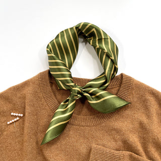 Moss green silk bandana with gold diagonal stripes tied around the neckline of a camel sweater, showcasing elegant styling with pearl pin accent