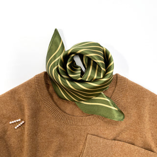 Moss green silk bandana with gold diagonal stripes elegantly tied around neck of camel sweater, showing luxurious sheen and modern maze pattern